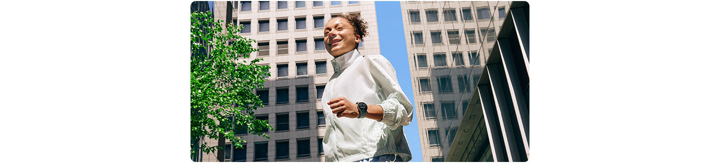 Uma mulher em uma roupa de ginástica vestindo um Galaxy Watch7 é vista andando em um quarteirão cercado por edifícios.