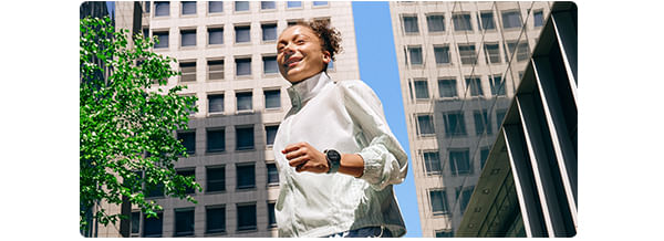 Uma mulher em uma roupa de ginástica vestindo um Galaxy Watch7 é vista andando em um quarteirão cercado por edifícios.